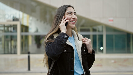 Wall Mural - Young beautiful hispanic woman student smiling confident talking on smartphone at street