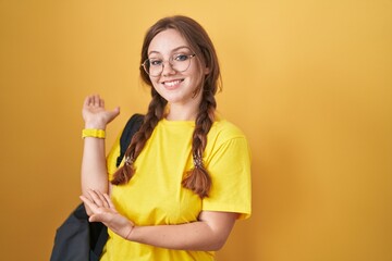 Wall Mural - Young caucasian woman wearing student backpack over yellow background inviting to enter smiling natural with open hand