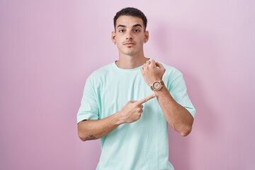 Poster - Handsome hispanic man standing over pink background in hurry pointing to watch time, impatience, looking at the camera with relaxed expression