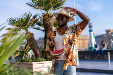 Wall Mural - Portrait of a black ethnic man enjoying summer vacation on the beach, wearing a hat and a watermelon smiling