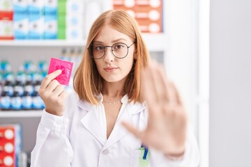 Poster - Young redhead woman working at pharmacy drugstore holding condom with open hand doing stop sign with serious and confident expression, defense gesture