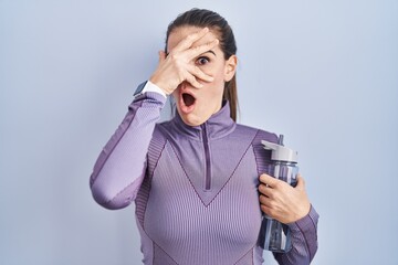 Poster - Beautiful woman wearing sportswear holding water bottle peeking in shock covering face and eyes with hand, looking through fingers with embarrassed expression.