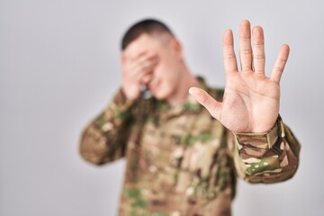 Wall Mural - Young man wearing camouflage army uniform covering eyes with hands and doing stop gesture with sad and fear expression. embarrassed and negative concept.