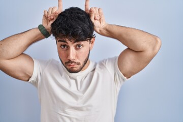 Sticker - Hispanic man with beard standing over white background doing funny gesture with finger over head as bull horns