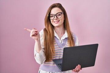 Canvas Print - Young caucasian woman working using computer laptop with a big smile on face, pointing with hand finger to the side looking at the camera.