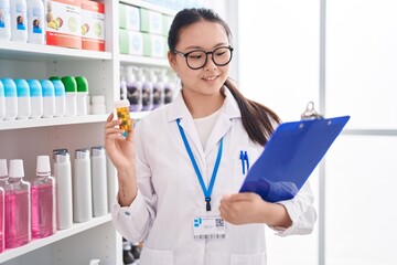 Canvas Print - Young chinese woman pharmacist holding pills bottle reading document at pharmacy