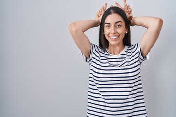 Wall Mural - Young brunette woman wearing striped t shirt posing funny and crazy with fingers on head as bunny ears, smiling cheerful