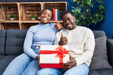 Sticker - Young african american couple holding present smiling happy and positive, thumb up doing excellent and approval sign