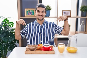 Wall Mural - Hispanic man with long hair sitting on the table having breakfast looking confident with smile on face, pointing oneself with fingers proud and happy.