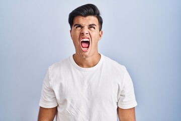 Sticker - Hispanic man standing over blue background angry and mad screaming frustrated and furious, shouting with anger. rage and aggressive concept.