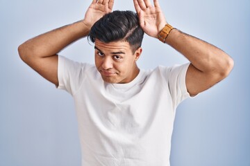 Poster - Hispanic man standing over blue background doing bunny ears gesture with hands palms looking cynical and skeptical. easter rabbit concept.