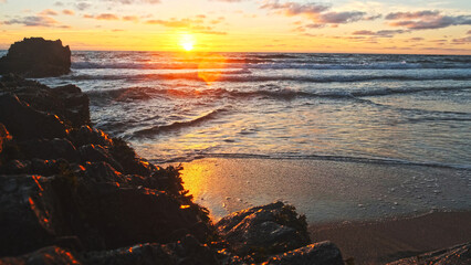 Poster - Beautiful natural landscape with sandy beach  with big stones during sunset.  Sun over horizon and Rolling ocean waves to the rocky shore while sunset.  Summer tourist destination in Portugal. Travel