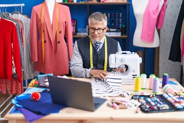 Poster - Middle age grey-haired man tailor using sewing machine and laptop at clothing factory