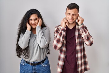 Poster - Young hispanic couple standing over white background covering ears with fingers with annoyed expression for the noise of loud music. deaf concept.