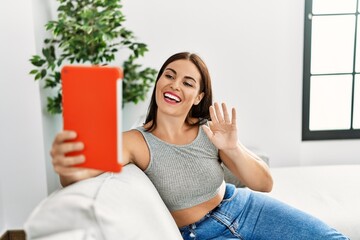 Wall Mural - Young beautiful hispanic woman having video call sitting on sofa at home