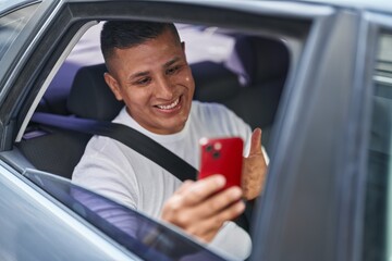 Poster - Young hispanic man doing video call with smartphone in the car smiling happy and positive, thumb up doing excellent and approval sign