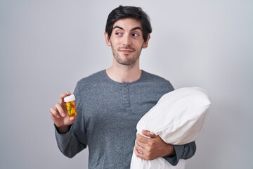 Canvas Print - Young hispanic man wearing pyjama hugging pillow holding pills smiling looking to the side and staring away thinking.