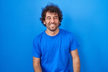 Poster - Hispanic young man standing over blue background winking looking at the camera with sexy expression, cheerful and happy face.