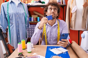 Young hispanic man tailor using smartphone and credit card with doubt expression at atelier