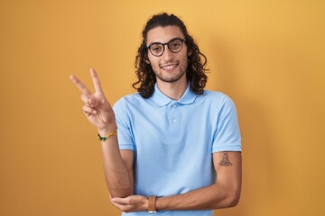 Poster - Young hispanic man standing over yellow background smiling with happy face winking at the camera doing victory sign with fingers. number two.