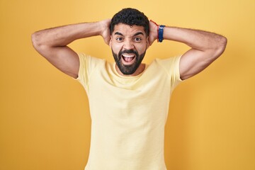 Sticker - Hispanic man with beard standing over yellow background crazy and scared with hands on head, afraid and surprised of shock with open mouth