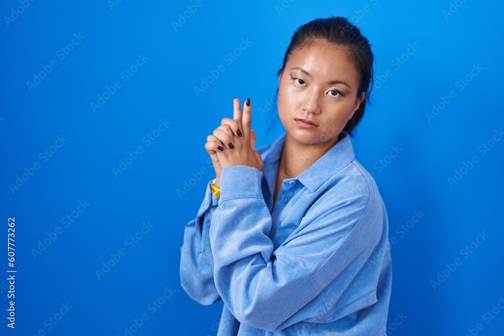 Asian young woman standing over blue background holding symbolic gun with hand gesture, playing killing shooting weapons, angry face
