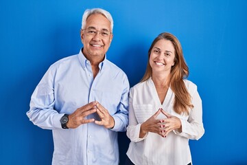 Wall Mural - Middle age hispanic couple standing over blue background hands together and fingers crossed smiling relaxed and cheerful. success and optimistic