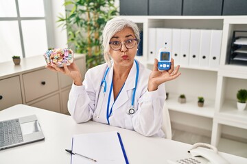 Canvas Print - Middle age woman with grey hair wearing doctor uniform holding glucose monitor puffing cheeks with funny face. mouth inflated with air, catching air.