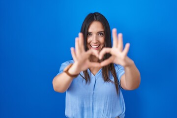 Canvas Print - Young brunette woman standing over blue background smiling in love doing heart symbol shape with hands. romantic concept.