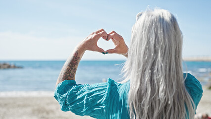 Poster - Middle age grey-haired woman doing heart gesture with hands at beach