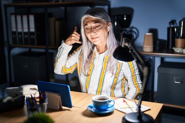 Poster - Middle age woman with grey hair working at the office at night smiling doing phone gesture with hand and fingers like talking on the telephone. communicating concepts.