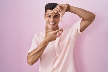 Sticker - Young hispanic man standing over pink background smiling making frame with hands and fingers with happy face. creativity and photography concept.