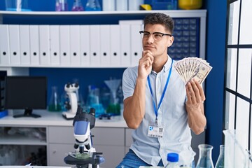 Sticker - Young hispanic man working at scientist laboratory holding dollars serious face thinking about question with hand on chin, thoughtful about confusing idea