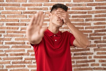 Canvas Print - Young hispanic man standing over bricks wall covering eyes with hands and doing stop gesture with sad and fear expression. embarrassed and negative concept.