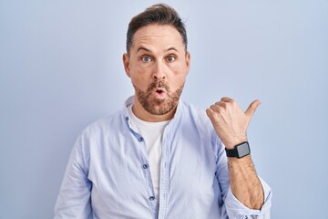 Poster - Middle age caucasian man standing over blue background surprised pointing with hand finger to the side, open mouth amazed expression.