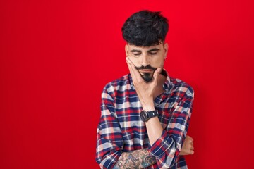 Wall Mural - Young hispanic man with beard standing over red background thinking looking tired and bored with depression problems with crossed arms.