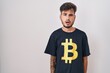 Young hispanic man with tattoos wearing bitcoin t shirt in shock face, looking skeptical and sarcastic, surprised with open mouth
