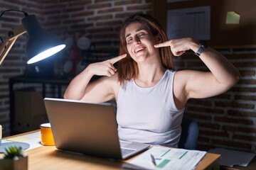 Wall Mural - Brunette woman working at the office at night smiling cheerful showing and pointing with fingers teeth and mouth. dental health concept.