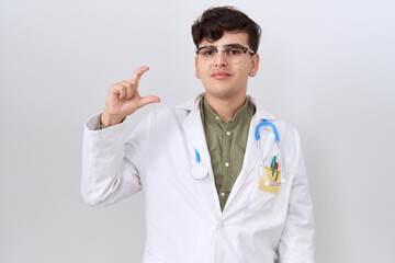 Sticker - Young non binary man wearing doctor uniform and stethoscope smiling and confident gesturing with hand doing small size sign with fingers looking and the camera. measure concept.