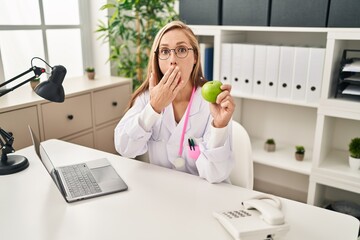 Sticker - Young blonde doctor woman holding green apple at the clinic covering mouth with hand, shocked and afraid for mistake. surprised expression