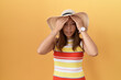 Middle age chinese woman wearing summer hat over yellow background suffering from headache desperate and stressed because pain and migraine. hands on head.