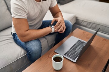 Wall Mural - Young hispanic man using laptop looking watch sitting on sofa at home