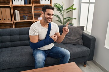 Canvas Print - Young hispanic man with beard wearing arm on sling sitting at therapy consult smiling happy pointing with hand and finger to the side