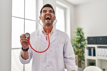 Poster - Young hispanic doctor man with beard holding stethoscope auscultating angry and mad screaming frustrated and furious, shouting with anger looking up.