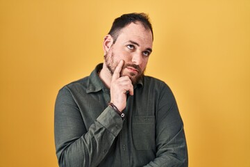 Wall Mural - Plus size hispanic man with beard standing over yellow background with hand on chin thinking about question, pensive expression. smiling with thoughtful face. doubt concept.