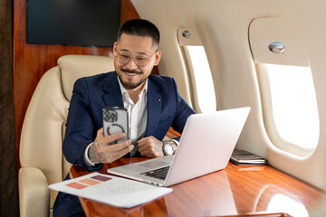 successful young asian businessman is sitting in private luxury jet with laptop and chatting via smartphone video call