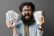 Handsome bearded man holds money in hand. Satisfied bearded man in denim shirt demonstrating bundle of money isolated on grey wall.