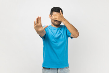 portrait of young adult unshaven man wearing blue t- shirt standing covering his face with hand, hid