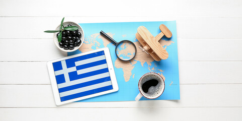 Wall Mural - Tablet computer with Greek flag on screen, world map, coffee, magnifier, olives and wooden airplane on white wooden background