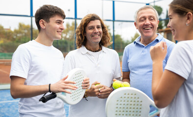 Wall Mural - Happy laughing men and woman of different ages in sportswear with rackets for padel and balls in hands talking friendly near net 